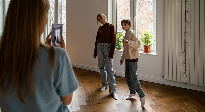 Kvinne i forgrunnen sett bakfra med mobilen, hun tar bilde av to barn som poserer foran et vindu i en ganske umøblert stue.